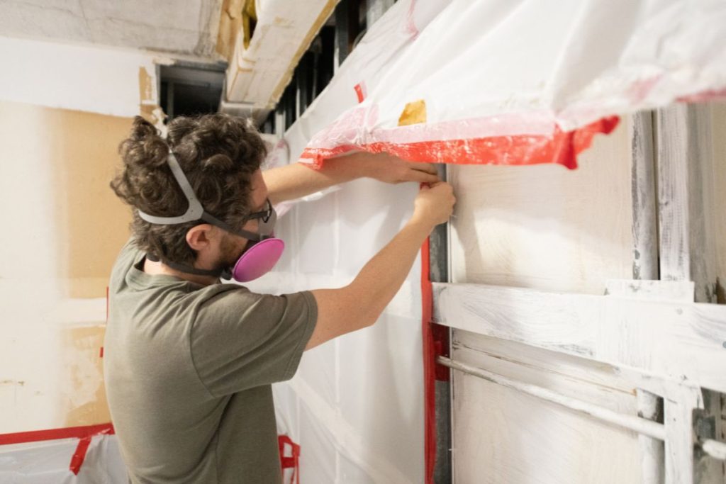 A masked and gloved man paints a wall as part of mold remediation procedures.