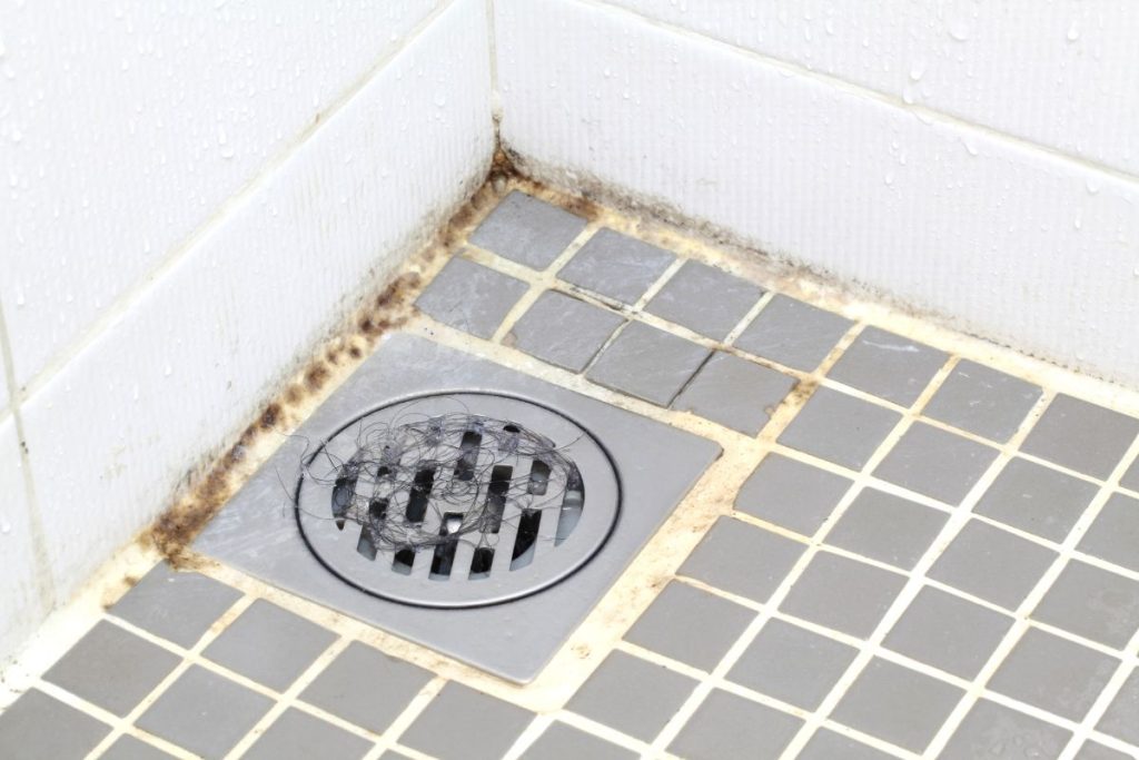 A shower drain set in a grimy tile floor, showing signs of dirt and neglect.